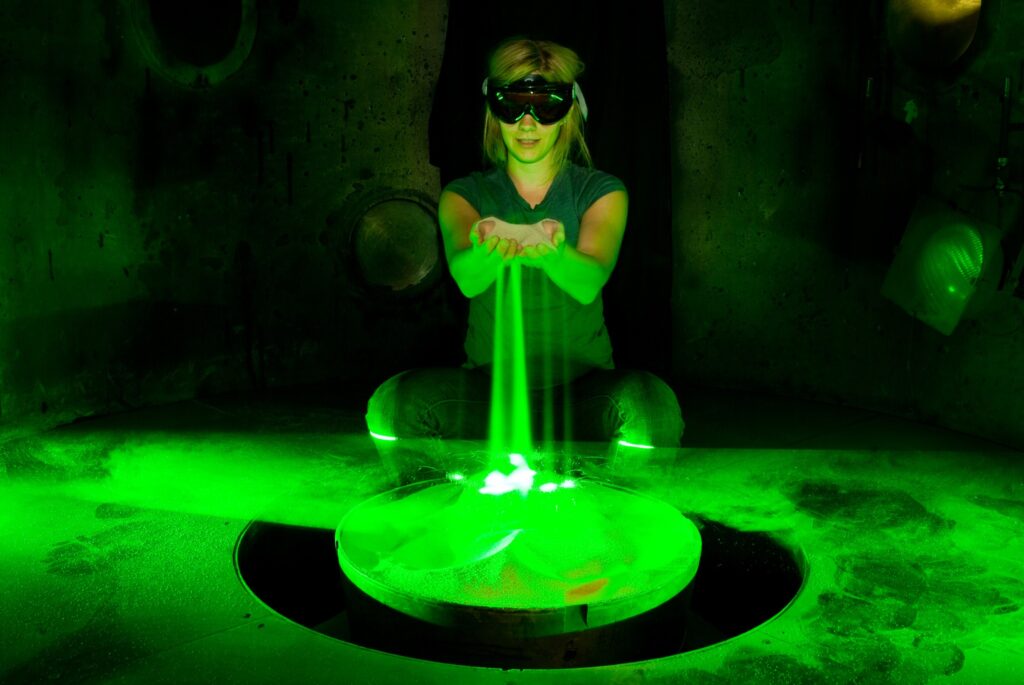 Sand glowing green from lasers slipping between hands of woman in dark room wearing goggles