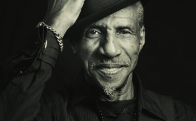 An older man wearing a dark hat and shirt smiles warmly while tilting his hat. He sports a goatee, a chain bracelet, and a beaded necklace.