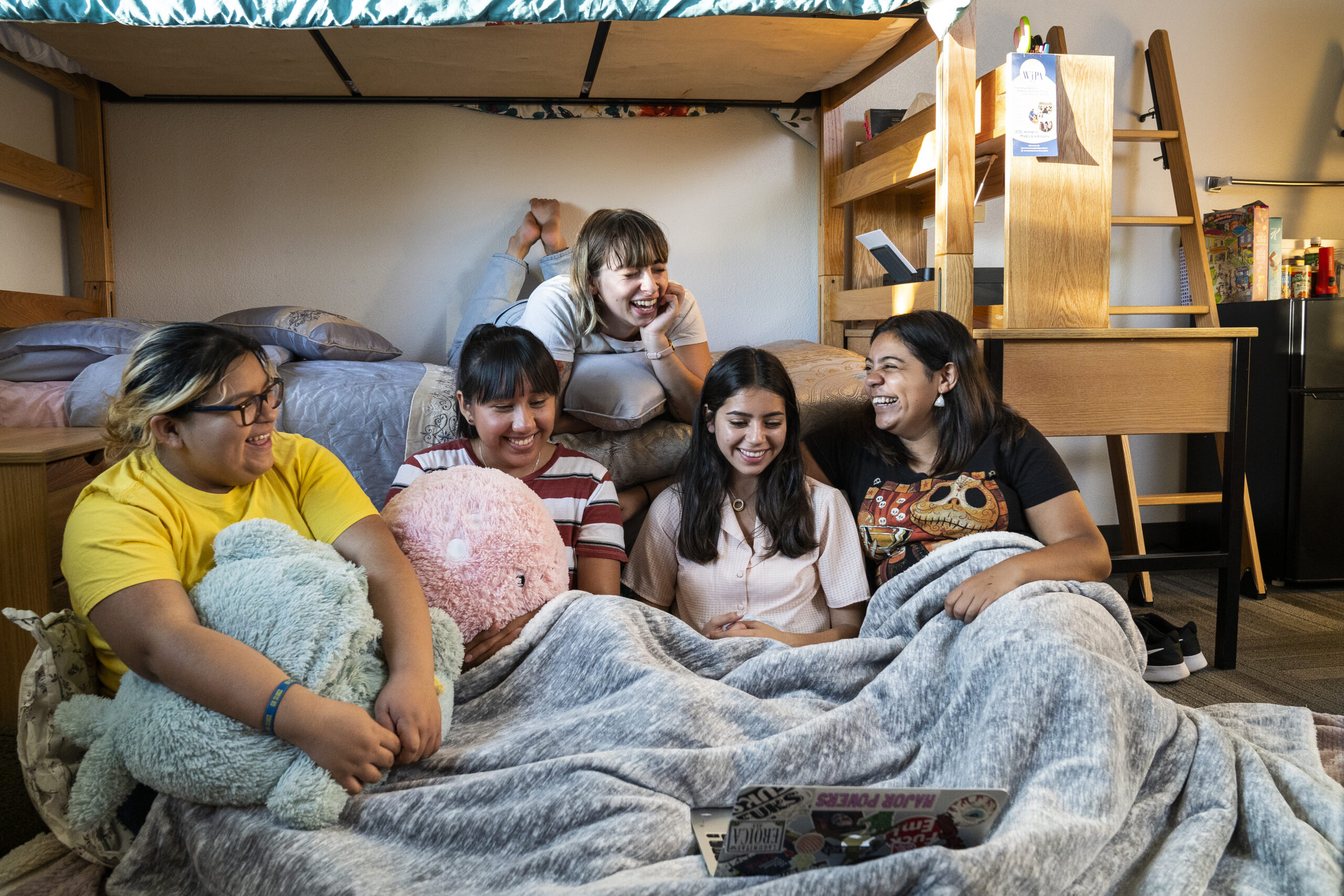 Five friends sit together in a dorm room, laughing and watching something on a laptop under a blanket. Plush toys and bunk beds surround them, creating a cozy atmosphere.