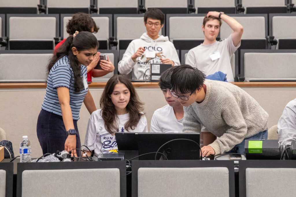 Four students in foreground, three in background, lean over computers 