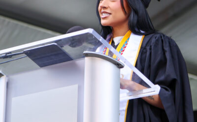 Becky G in cap and gown speaks at a podium under a large tent. She wears a medal and stole while delivering her speech.