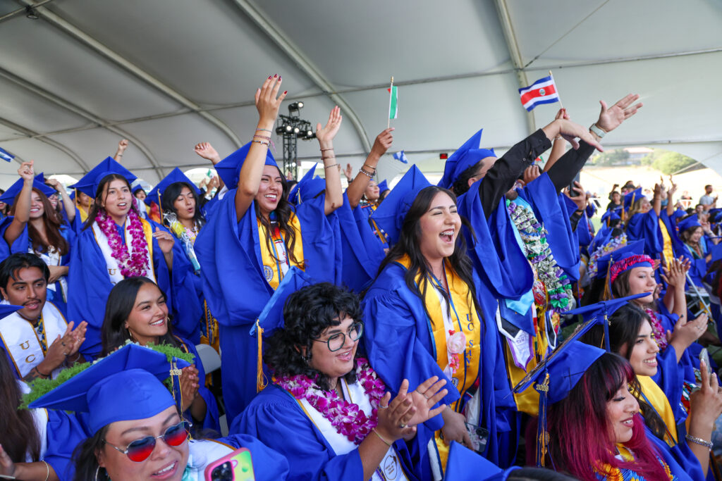 Graduates in blue caps and gowns cheer and celebrate under a large tent, some waving flags and wearing leis. The crowd is smiling, clapping, and raising their hands in excitement.