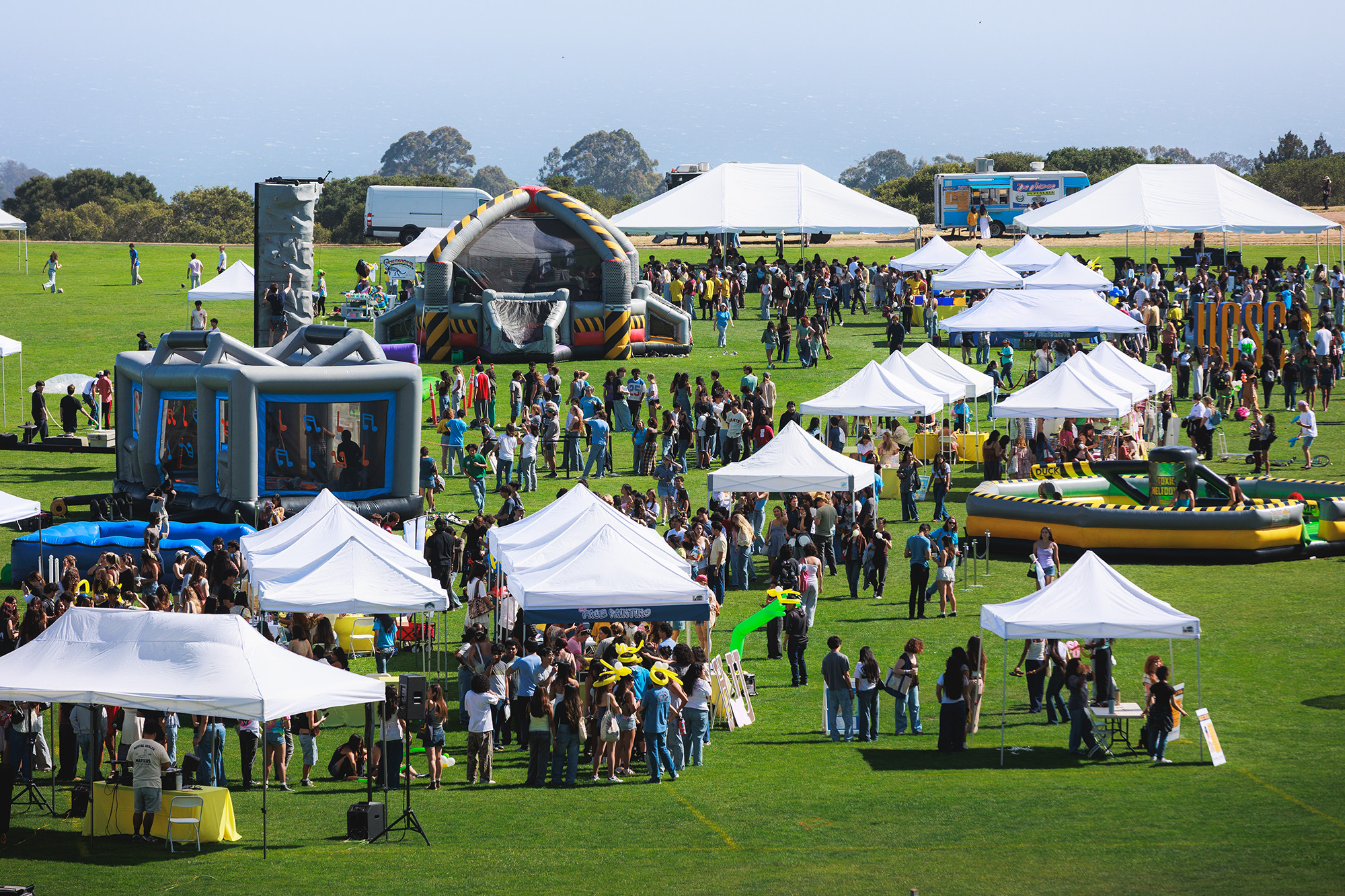 An outdoor daytime aerial view of the bustling Carnival on UCSC's East Field. Numerous white canopy tents are scattered across the lawn, with crowds of people walking between them, queuing in lines, and gathering in groups. The event features large inflatable attractions, including a gray and yellow obstacle course on the left and a round, yellow and black "8-Player Meltdown" game on the right.