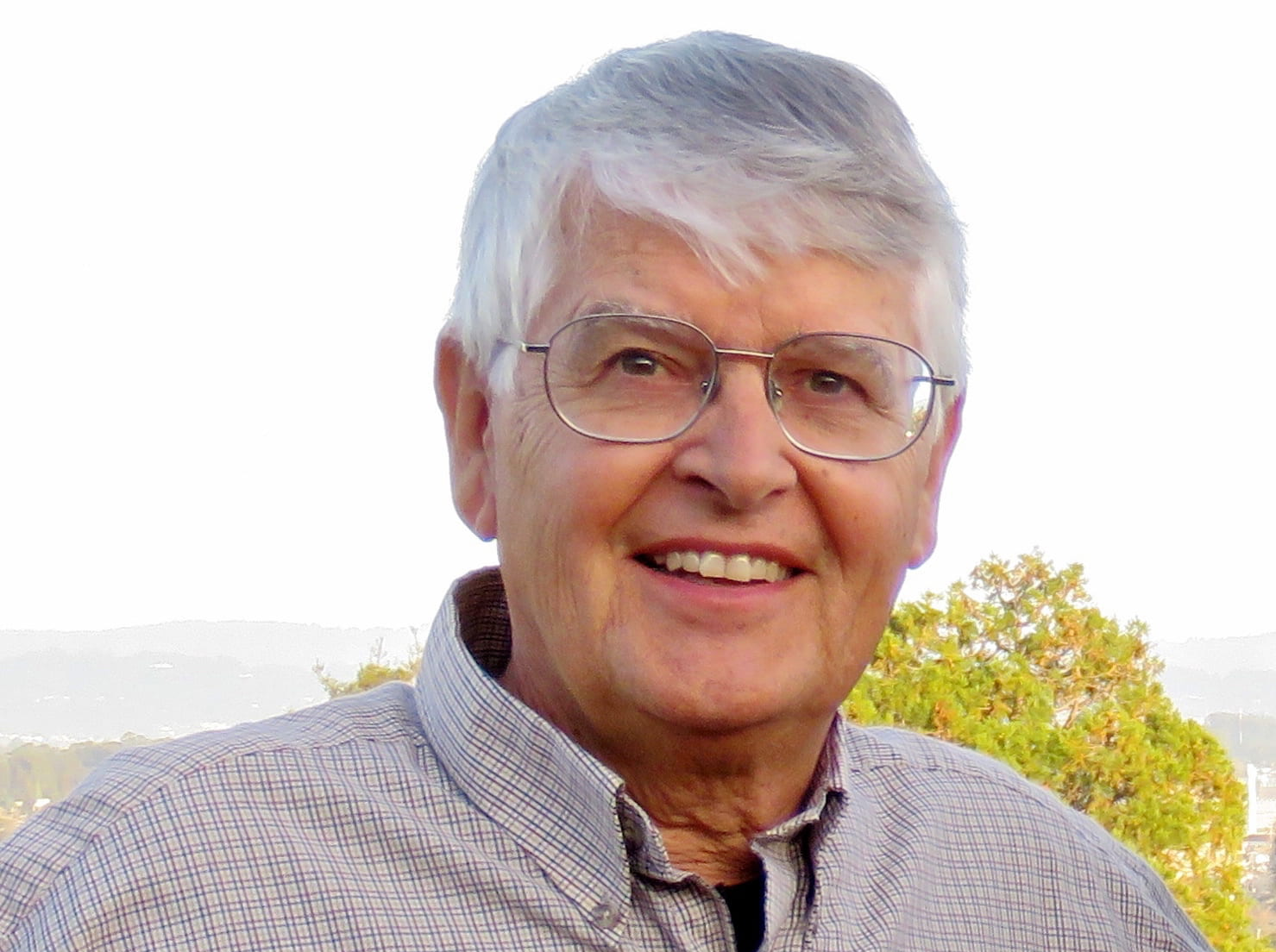 An older man with gray hair and glasses is smiling outdoors. He is wearing a light-colored, checkered shirt with trees and a pale sky in the background.
