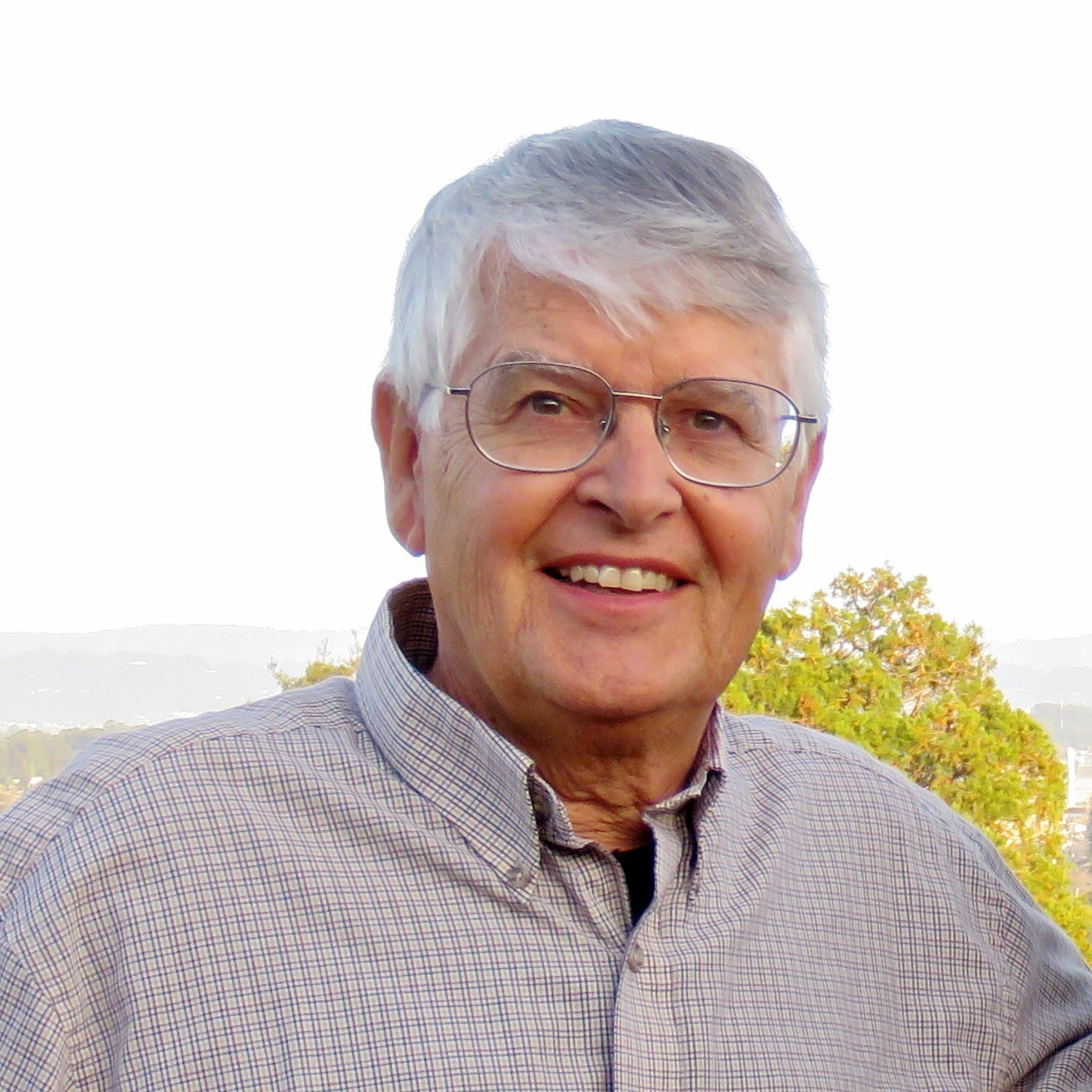 An older man with gray hair and glasses is smiling outdoors. He is wearing a light-colored, checkered shirt with trees and a pale sky in the background.