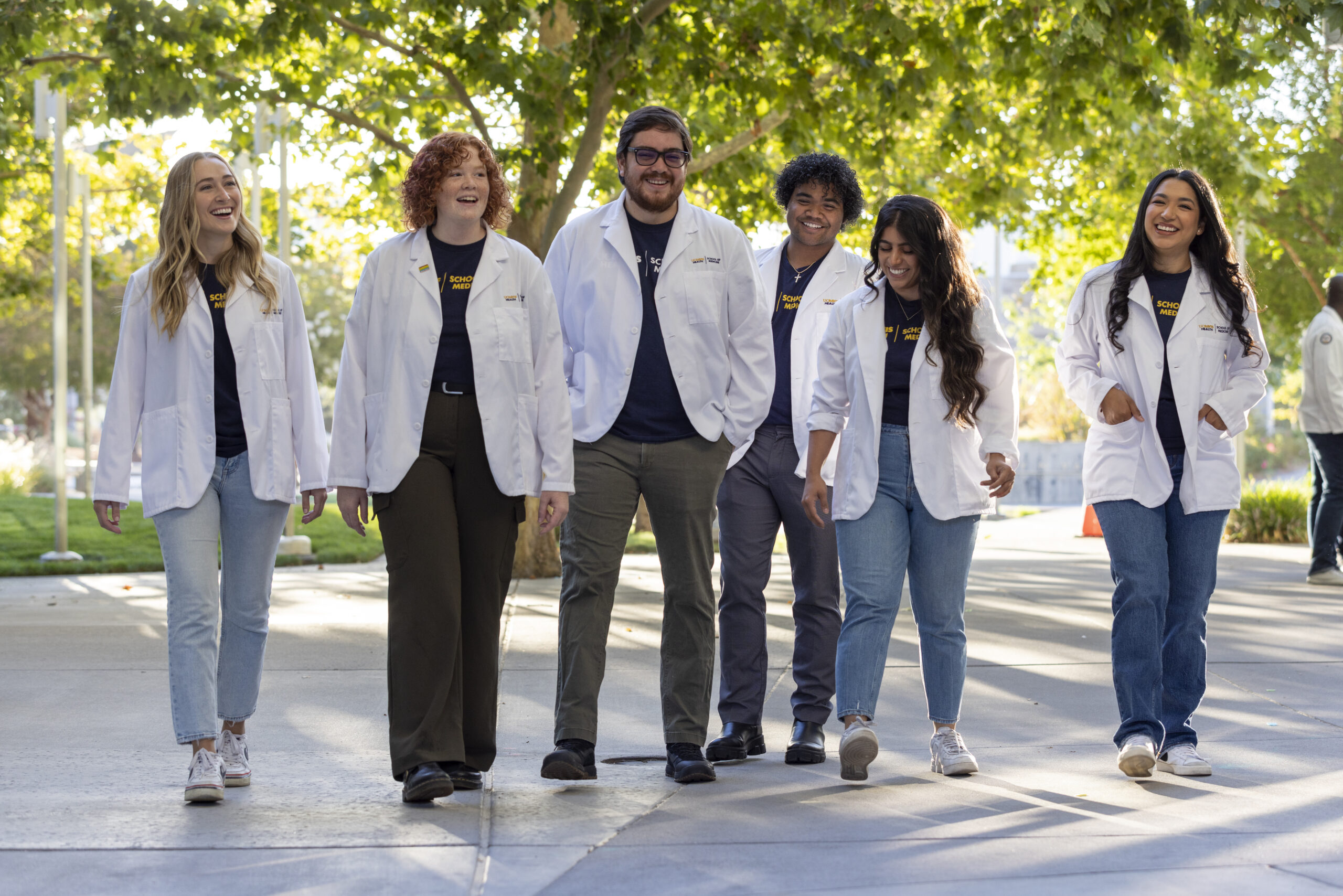 Six smiling students in white lab coats walk outdoors under green trees. They appear relaxed and cheerful.