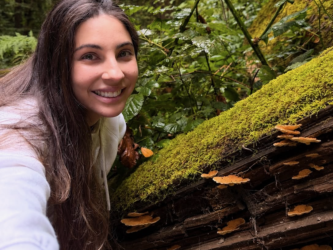 A young woman leans over a sideways tree trunk that mushrooms are growing out of