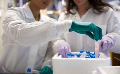 In focus is a hand pulling a vial out of a container. Two figures in white lab coats can be seen behind.