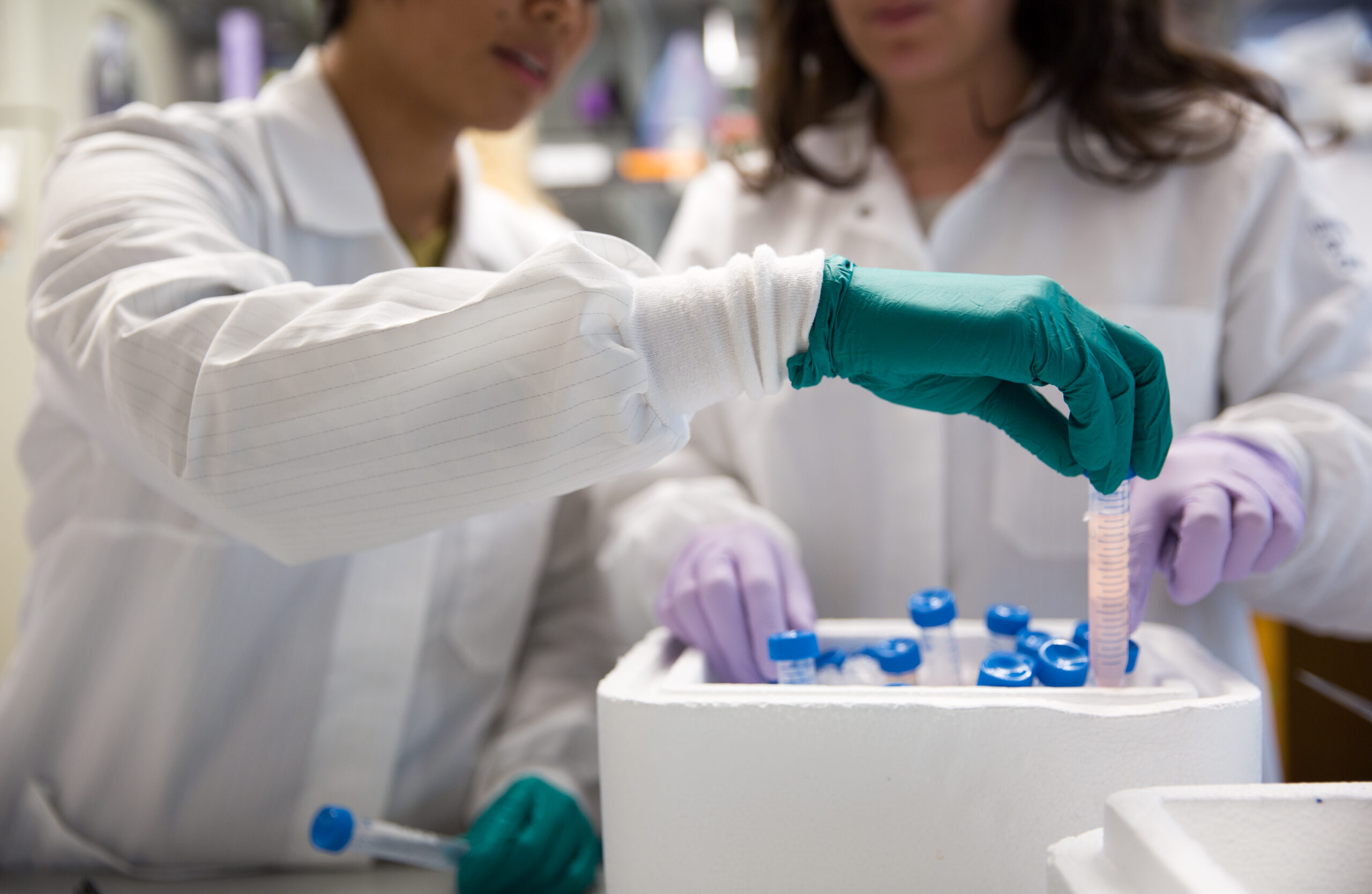 In focus is a hand pulling a vial out of a container. Two figures in white lab coats can be seen behind.