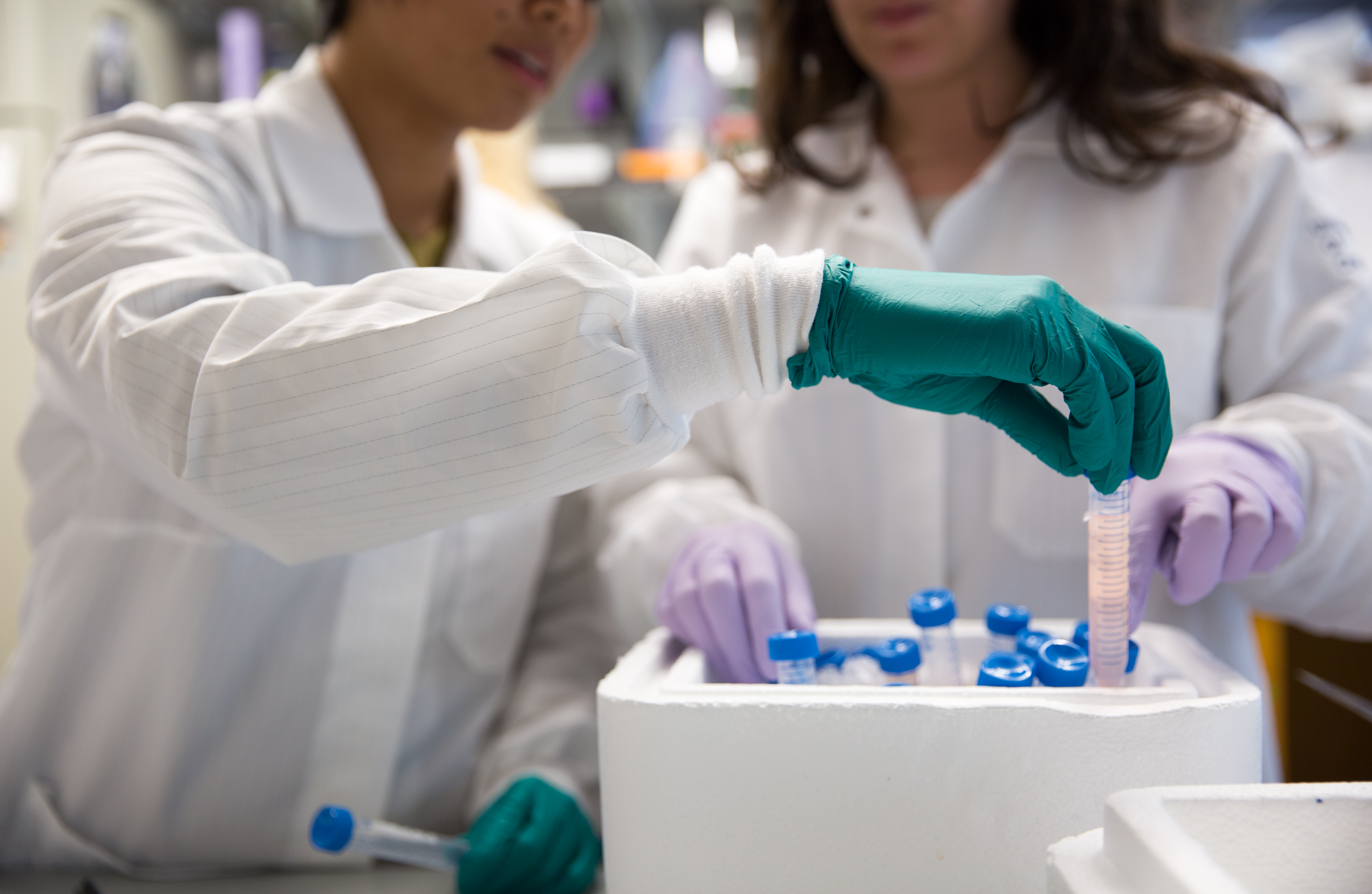 In focus is a hand pulling a vial out of a container. Two figures in white lab coats can be seen behind.