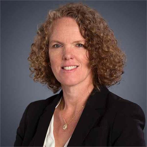 A woman with curly, shoulder-length hair is wearing a black blazer over a white top. She is smiling slightly against a dark gray background.