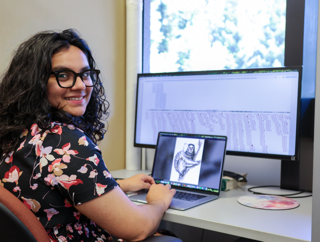 A woman sits at a desk in front of a computer displaying an image of a marmoset and data related to genomics.