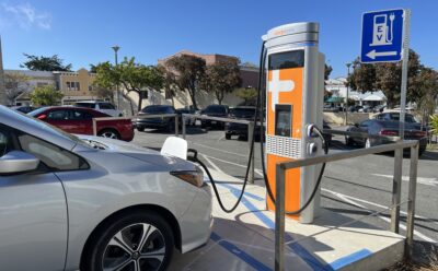 Car plugged in to a charger in a parking lot of a downtown area, with an "EV" sign.