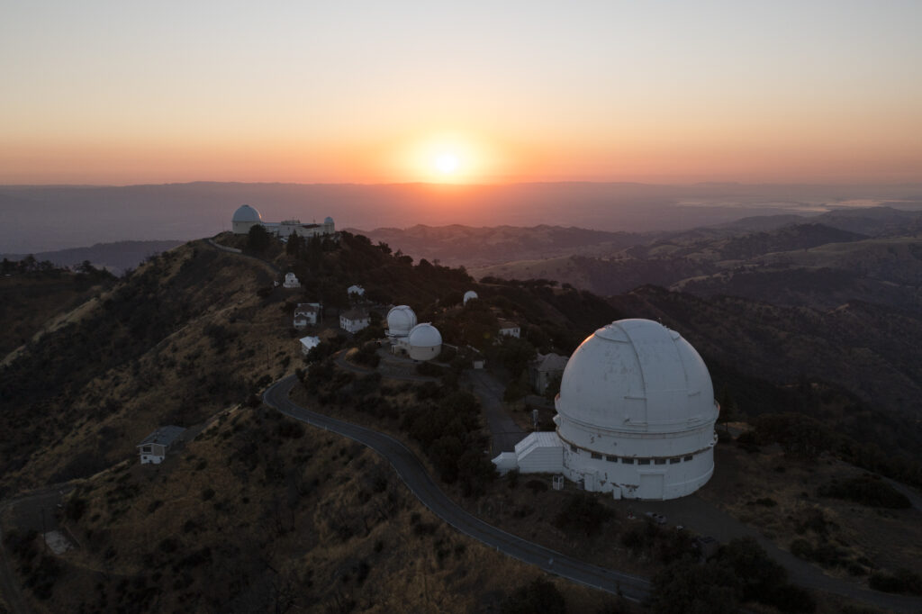 Sunset illuminates a mountain ridge dotted with observatories and winding roads. The large white domes contrast against the darkening landscape.