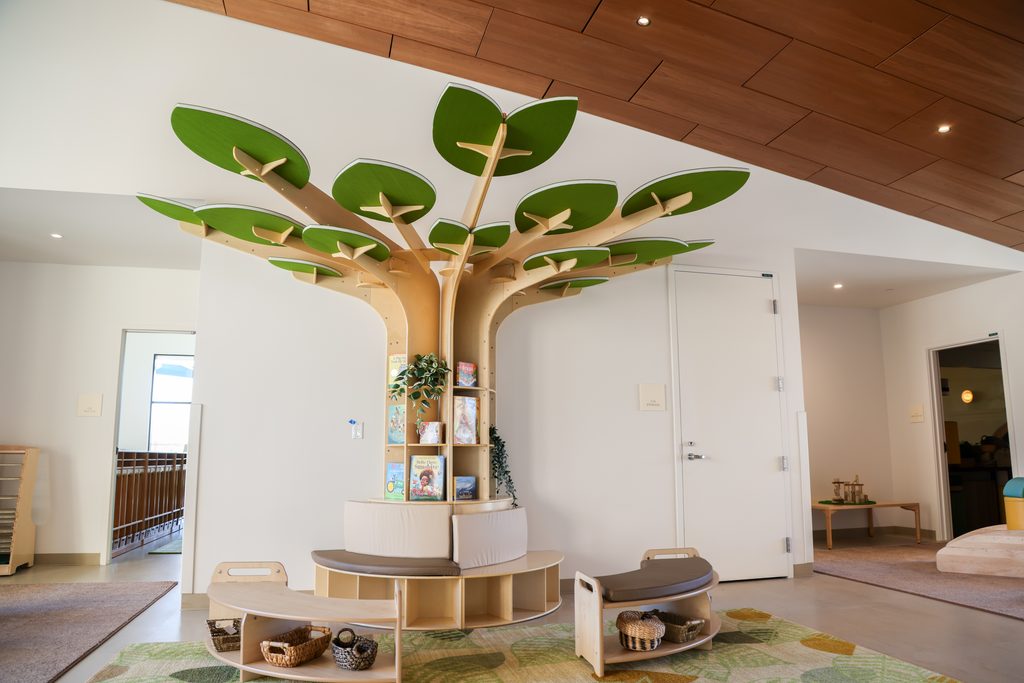 Interior view of the new Early Education Center showing a large wooden tree-shaped bookshelf surrounded by child-size seating on a cozy rug