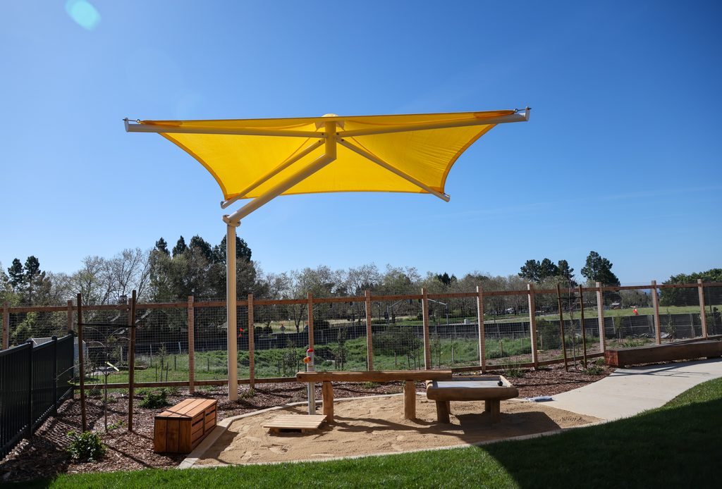 View of an outdoor play area at the new UCSC Early Education Center showing a yellow shade covering, wooden benches, and play table