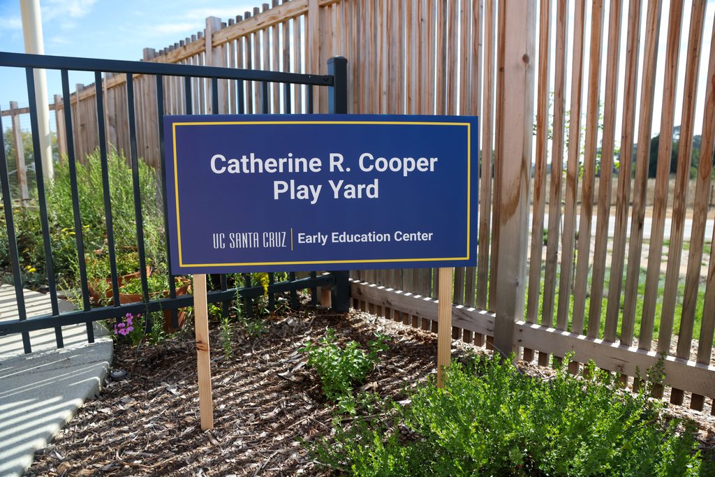 A blue yard sign reading "Catherine R. Cooper Play Yard – UC Santa Cruz Early Education Center" staked into a garden bed beside a play yard fence