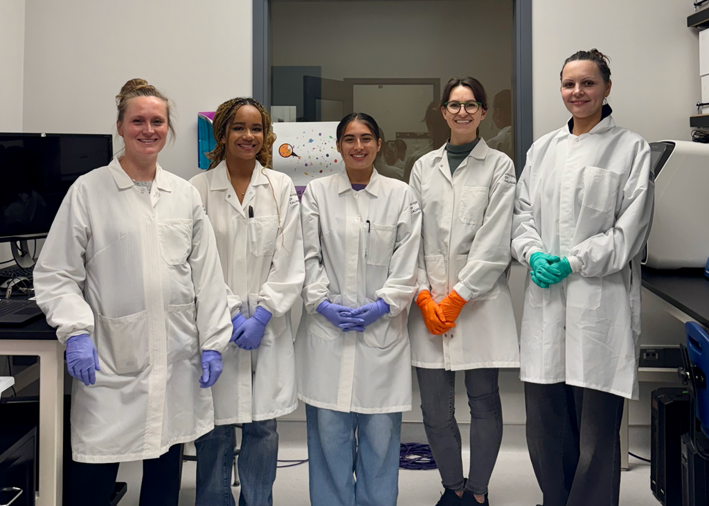 Five women in lab coats and gloves in a laboratory space.