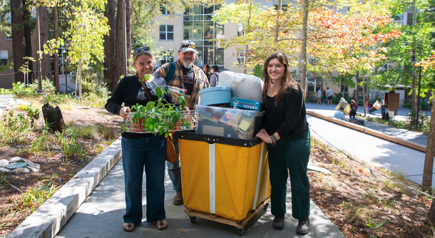 Three people stand beside a large move-in cart filled with bins, boxes and a potted plant on the UC Santa Cruz campus.
