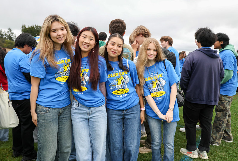 Four students wearing matching blue Slug Life T-shirts pose together on a field during a UC Santa Cruz welcome event, with a crowd of students behind them.