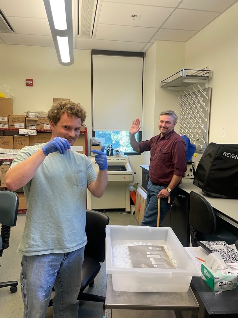 Lab researcher in foreground holding sample; second researcher smiling and waving in the background