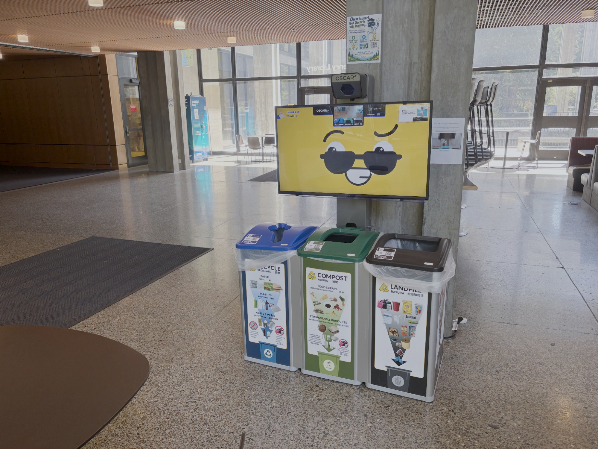 A waste station with bins for recycling, compost, and landfill sits in a bright, open lobby. Above the bins, a screen with a smiling face and sunglasses is mounted on a concrete pillar.