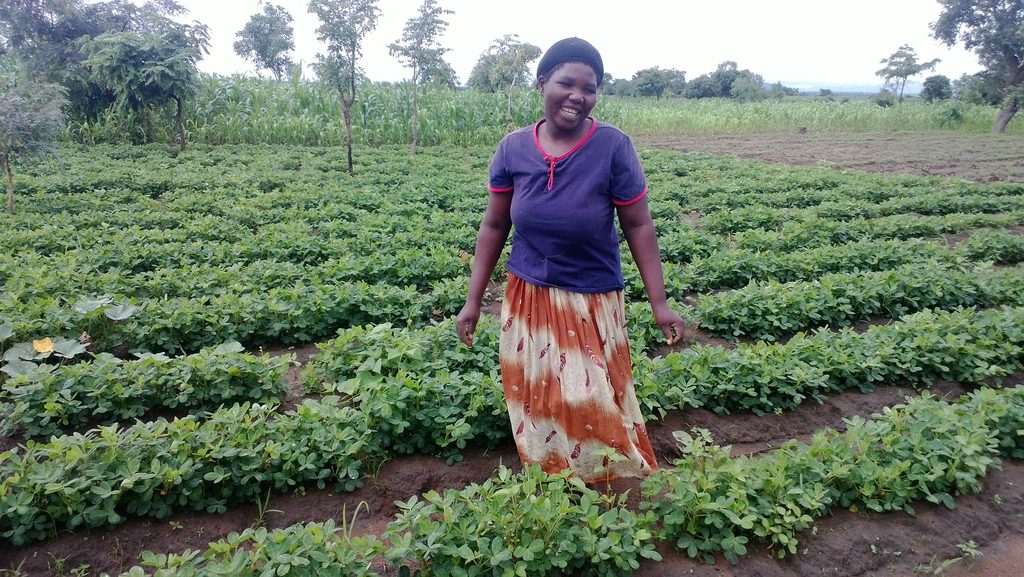 A woman smiles as she stands amidst rows of crops in a farm field.