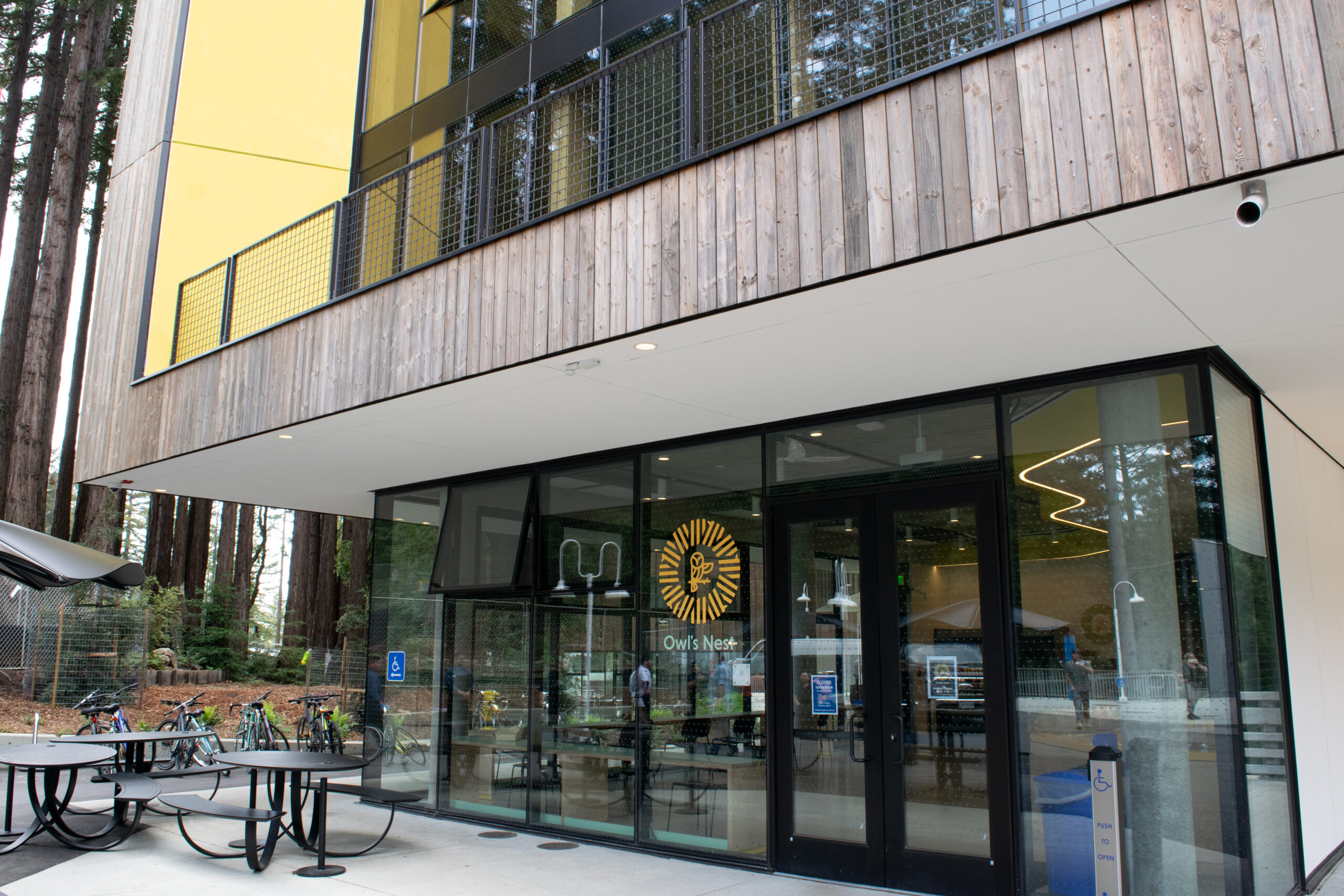 Exterior of a modern building surrounded by tall redwood trees, featuring wood siding, bright yellow wall panels, large glass windows, and black metal railings. The ground-floor entrance is labeled “Owl’s Nest,” with outdoor picnic tables, bicycles, and accessible-entry signage in front.