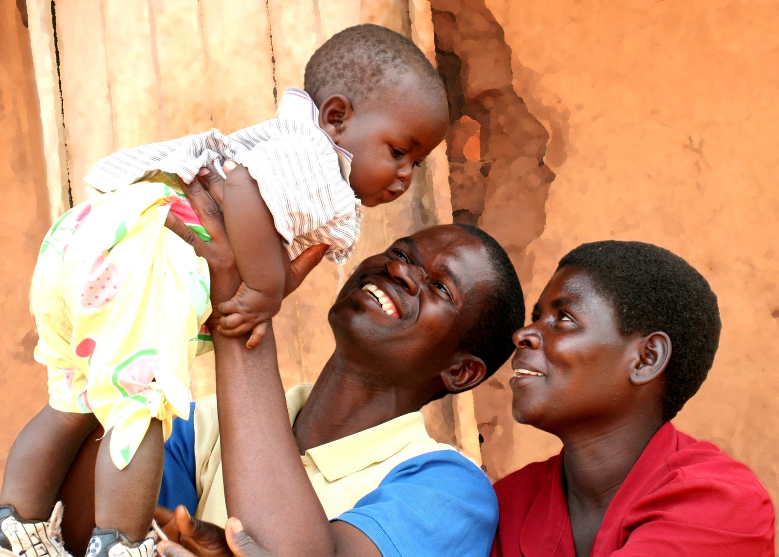 A father lifts his baby daughter as father and mother sit side by side outside a building in Malawi and smile at the baby.