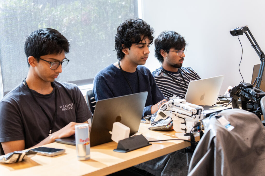 Three students work with laptop computers and a robotic arm