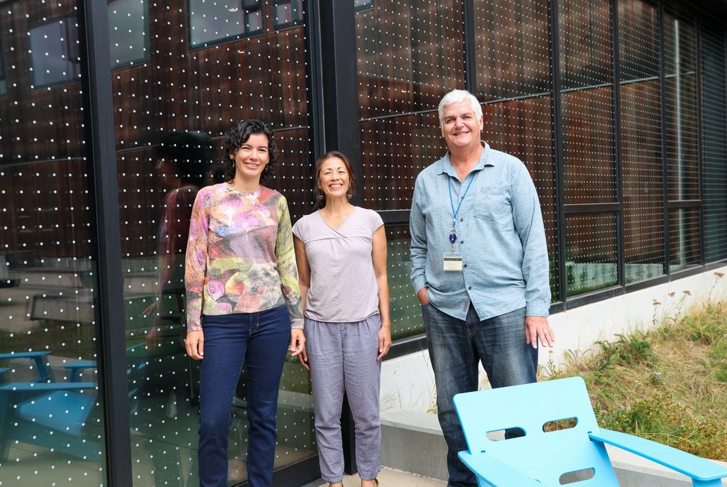Three people stand in front of a set of large windows, with dot window film showing. 